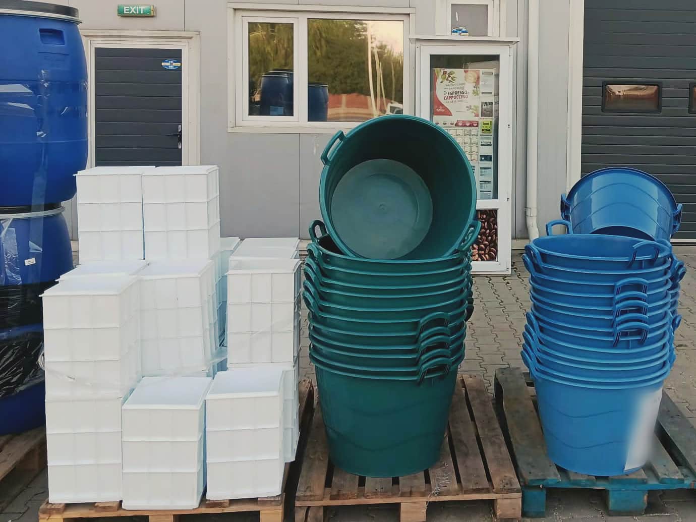 Lighene mari de plastic verzi și albastre, lăzi albe și un butoi albastru, pe paleți în fața magazinului