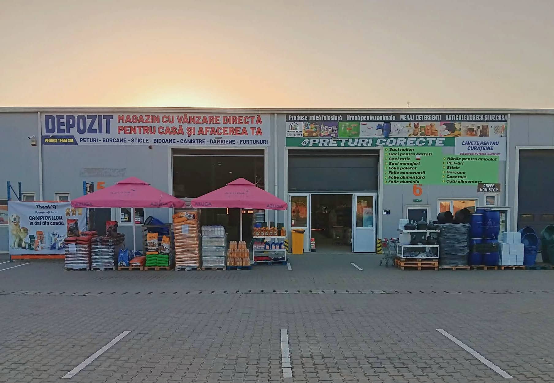 Fațada magazinului Pedros Team la apus: firma DEPOZIT cu litere albastre, panoul verde „Prețuri corecte”, două umbrele roz și paleți cu marfă în fața halei